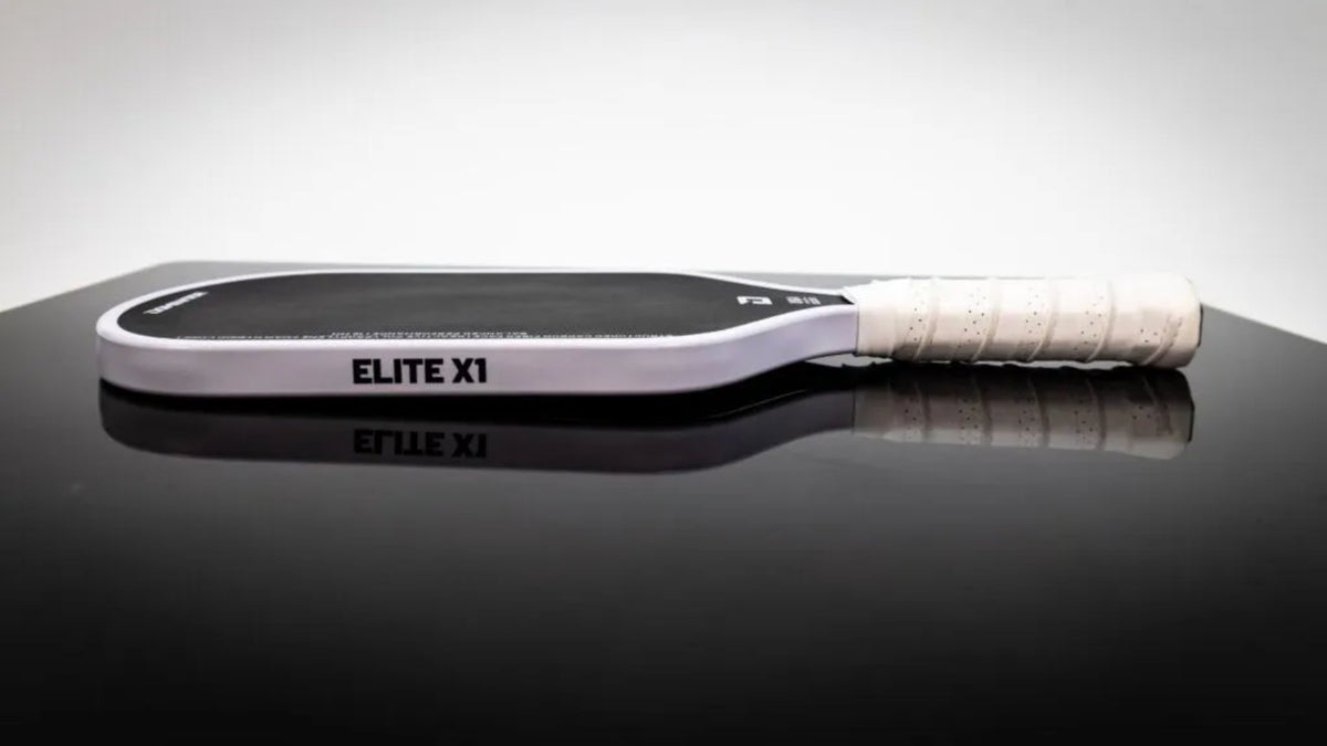 A pickleball paddle with a black grip, white frame, and white wrapped handle, resting on a reflective black surface against a white background. The brand name 'ELITE XI' is printed on the paddle face.