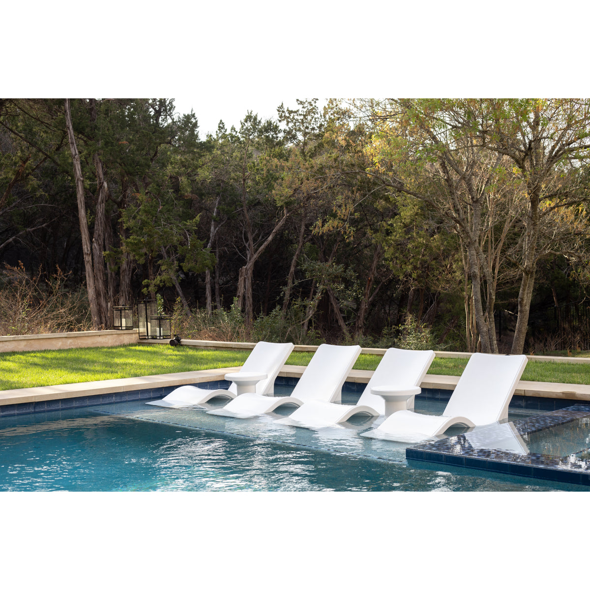 Row of four white baja shelf lounge chairs on an in-pool sun shelf with a matching floating side table between them, backed by mature trees and a grassy lawn, SKU 8902-700 in Highcloud White.