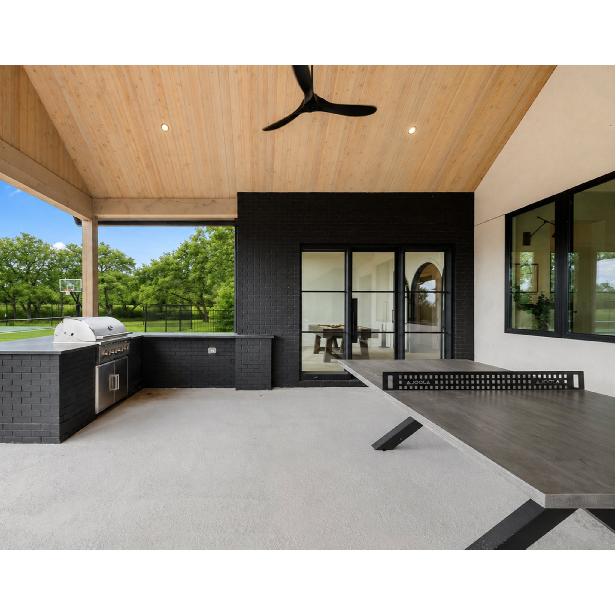 Lifestyle shot of the JOOLA outdoor ping pong table set up on a covered patio beside a black brick outdoor kitchen with a built-in grill, framed by an open-beam wood ceiling and lawn views, SKU 11120-11121, Gray.