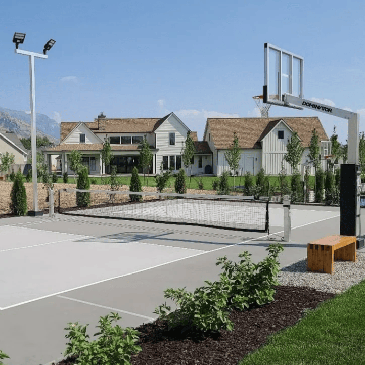 Outdoor multi-sport court in a residential neighborhood with a pickleball net, basketball hoop, court lighting, painted markings, landscaping, nearby homes, and mountains in the background. Learn how to plan an entertainment space around your available area, preferred activities, amenity mix, layout, and how people will gather and use the space.